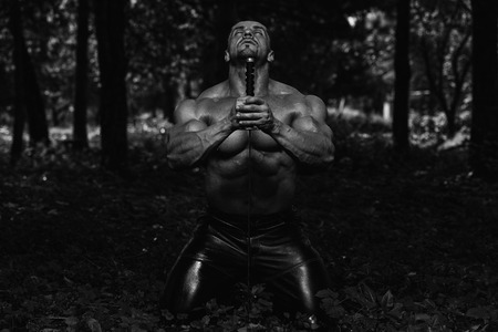 Action Hero Muscled Man Holding A Ancient Sword - Standing In Forest Wearing Leather Pantsの写真素材
