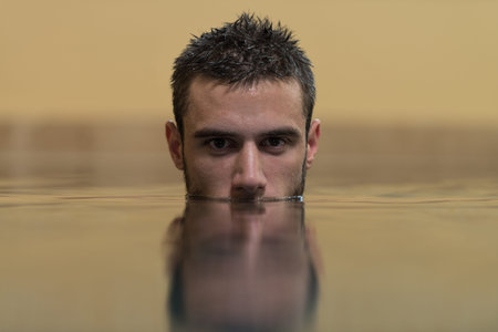Young Man Swimming In Pool With Face Half Submerged Looking At Camera