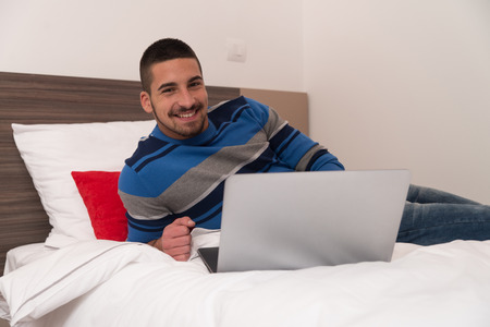 Young Male Student Lying On Bed And Having Fun With Laptop In Bedroomの写真素材