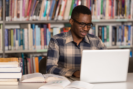 In The Library - Handsome African Male Student With Laptop And Books Working In A High School - University Library - Shallow Depth Of Fieldの写真素材