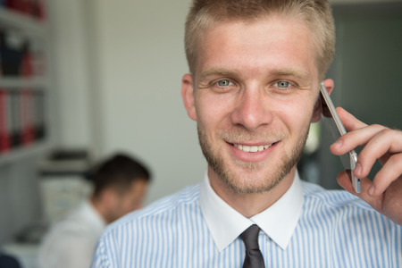 Happy Young Business Man Talking On Telephone In Modern Office Behind Him Sitting His Colleagueの写真素材