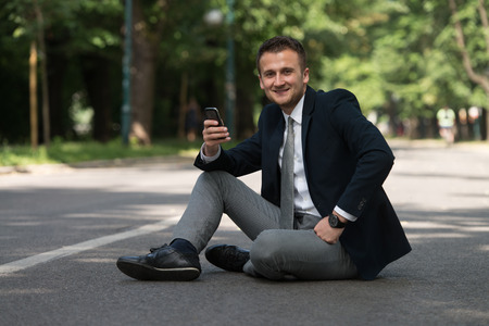 Happy Business Man Using Mobile Phone Outside Sitting On Asphaltの写真素材
