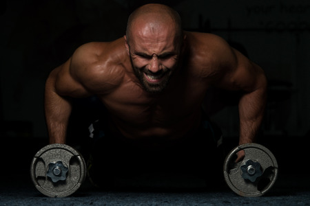 Young Adult Athlete Doing Push Ups With Dumbbells As Part Of Bodybuilding Training In A Dark Roomの写真素材