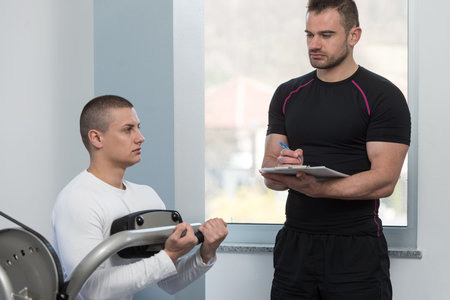 Personal Trainer Working With A Young Man At The Gym Writing Notes On A Clipboard In A Health And Fitness Conceptの写真素材