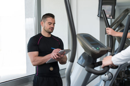 Group Of People Exercising On Elliptical Walker In Gym Or Fitness Club While Personal Trainer With...