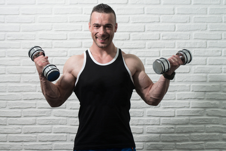 Young Man Working Out Biceps With Dumbbells On White Bricks Background With Copyspace - Dumbbell Concentration Curlsの写真素材