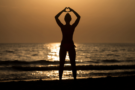 Beautiful Female Model Enjoying Sunset and Making Heart Sign on Sun at Seaside - Calm Water Reflects Silhouette of Woman - Sun Goes Under Horizonの写真素材