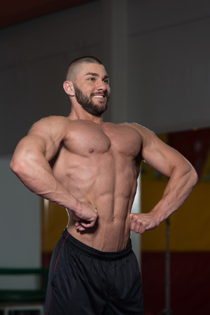 Healthy Young Man Standing Strong In The Gym And Flexing Muscles - Muscular Athletic Bodybuilder Fitness Model Posing After Exercisesの写真素材