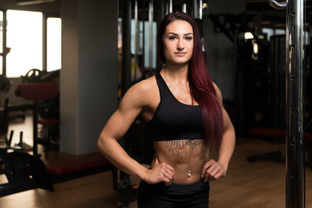 Healthy Young Woman Standing Strong In The Gym And Flexing Muscles - Beautiful Athletic Fitness Model Posing After Exercisesの写真素材