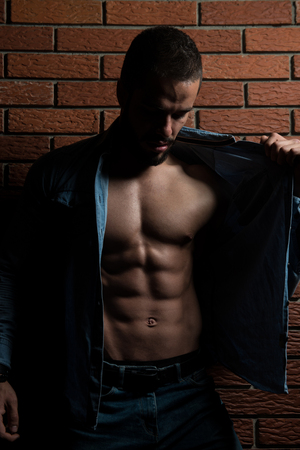 Healthy Young Man Standing Strong In The Gym And Flexing Muscles While Wearing Blue Shirt - Muscular Athletic Bodybuilder Fitness Model Posing After Exercises On Wall of Bricksの写真素材