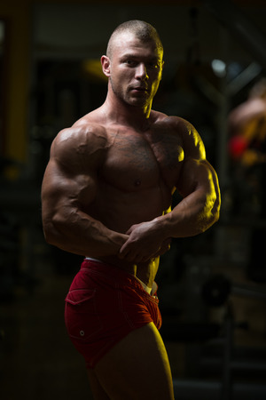 Handsome Young Man Standing Strong In The Gym And Flexing Muscles - Muscular Athletic Bodybuilder Fitness Model Posing After Exercisesの写真素材