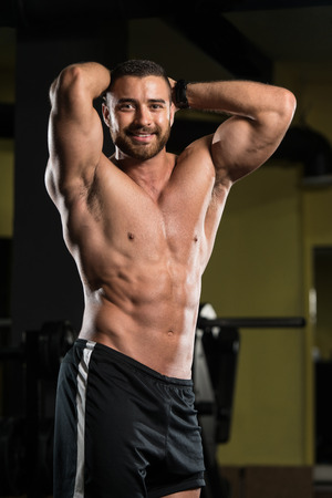 Handsome Young Man Standing Strong In The Gym And Flexing Muscles - Muscular Athletic Bodybuilder Fitness Model Posing After Exercisesの写真素材