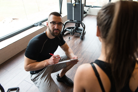 Personal Trainer Takes Notes While Young Woman Resting On Bench In Gymの写真素材