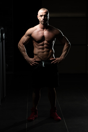 Handsome Young Man Standing Strong In The Gym And Flexing Muscles - Muscular Athletic Bodybuilder Fitness Model Posing After Exercisesの写真素材
