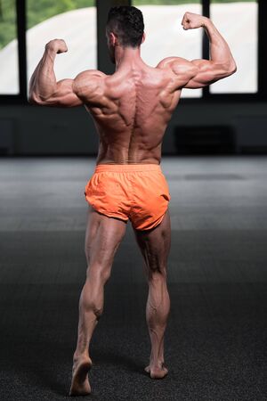 Handsome Young Man Standing Strong In The Gym And Flexing Muscles - Muscular Athletic Bodybuilder Fitness Model Posing After Exercisesの写真素材