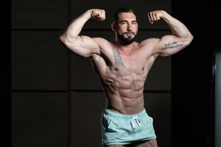 Young Man Standing Strong In The Gym And Flexing Muscles - Muscular Athletic Bodybuilder Fitness Model Posing After Exercisesの写真素材