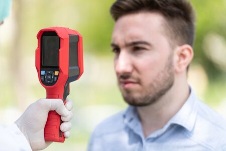 Doctor Man Use Infrared Forehead Thermometer Gun to Check Body Temperature for Virus Covid-19 Symptoms With the Isolation Gown or Protective Suits and Surgical Face Masks Outdoors in Parkの写真素材