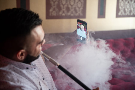 Young Man Smoking Shisha At Arabic Restaurant - Man Exhaling Smoke Inhaling From A Hookah and Taking Selfie With Phoneの写真素材