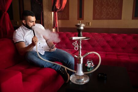 Man Smoking Turkish Hookah In The Cafe With Colorful Walls On Background and Texting on Phoneの写真素材