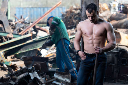 Handsome Man Standing Strong in Scrap Metal Industrial Junkyard and Flexing MusclesW ith Shovel - Muscular Athletic Bodybuilder Fitness Model Posingの写真素材