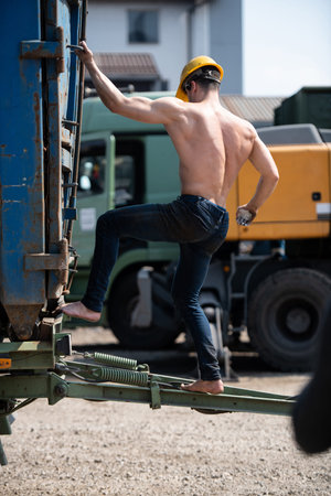 Young Man Athlete is Climbing Onto the Trailer Outdoors in Industrial Yard and Wearing Yellow Helmet