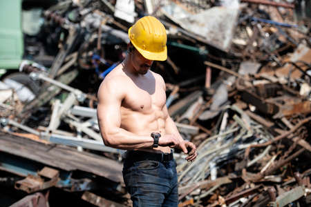 Handsome Man Standing Strong in Scrap Metal Industrial Junkyard and Flexing Muscles - Muscular Athletic Bodybuilder Fitness Model Posing With Yellow Helmetの写真素材