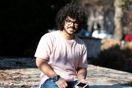 Portrait of Handsome Young Afro Man Talking Touching Phone or Using Smartphone for the Call and Sitting Outside During Autumnの写真素材