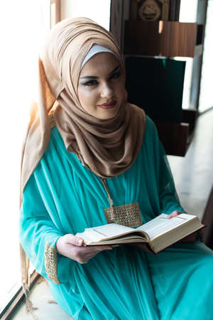 Beautiful Muslim Woman Sitting on Knees Reading the Quran and Turning Page on the Bookの写真素材