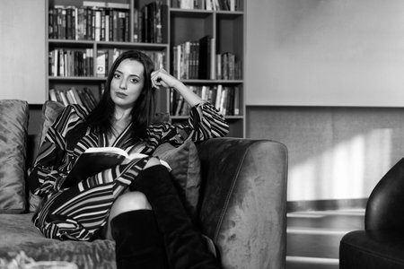Pretty Woman With Black Hair Sitting on Couch in the Library - Blurred Books at the Backの写真素材