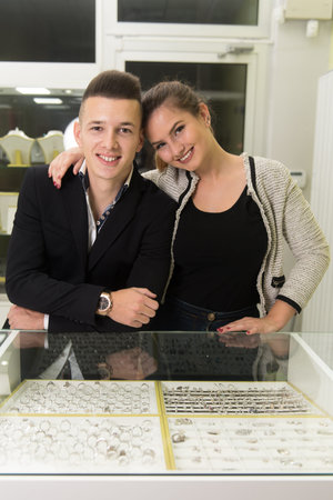 Attractive Young Couple Inspecting Selection of Rings While Her Loving Boyfriend Standing by at the...