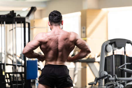 Handsome Young Man Standing Strong In The Gym And Flexing Muscles - Muscular Athletic Bodybuilder Fitness Model Posing After Exercisesの写真素材