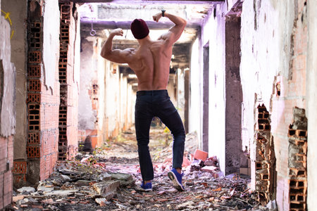Handsome Young Man With Winter Hat Standing Strong Outdoors and Flexing Back Muscles in Ruins - Muscular Athletic Bodybuilder Man Posingの写真素材