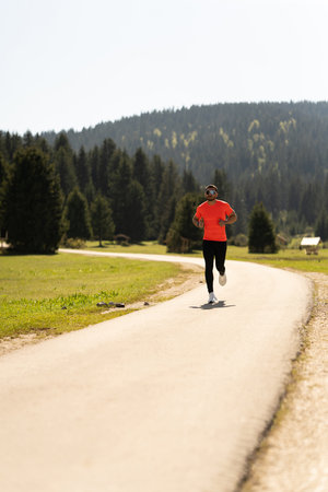 Young Man Runs On A Scenic Outdoor Pathway Surrounded By Grass, Trees, And Mountains On A Sunny Day In Natureの写真素材