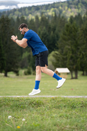 Man In Sporty Outfit Stands Focused And Ready For A Sprint, Framed By Green Grass, Trees, And A Mountain Viewの写真素材