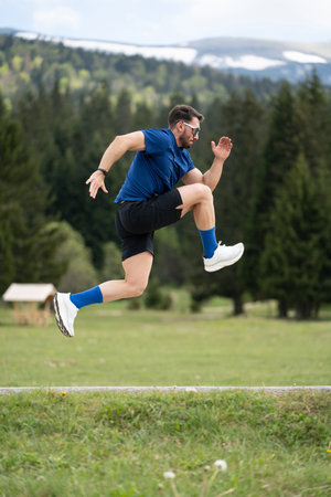 Man In Sporty Outfit Stands Focused And Ready For A Sprint, Framed By Green Grass, Trees, And A Mountain Viewの写真素材