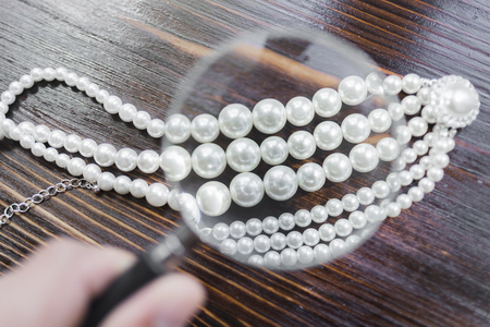 Pearl necklace with a large white pearl surrounded by diamonds under a magnifying glass on a dark brown background.の写真素材