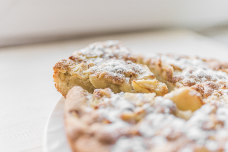 A fresh baked apple pie and a cut piece are sprinkled with sugar powder and lie on a white plate on a light background.の写真素材