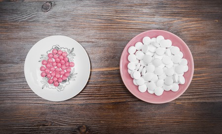 Bright pink pills on a white saucer and big white pills on a pink saucer on a wooden dark backgroundの写真素材