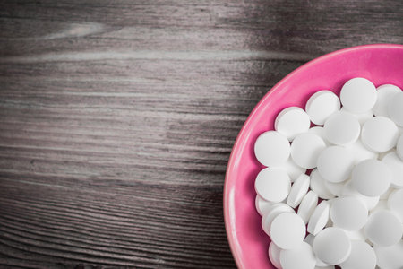 Many large white pills on a small bright pink plate on a brown wooden background, on the right side...