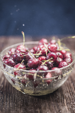 Juicy ripe sweet cherry lies in a transparent bowl under the spray of drops of water on a dark brown wooden background.の写真素材