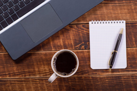 Laptop or notebook with cup of coffee and notepad with pen on old wooden table background.