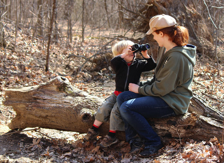 Blond boy looking through binoculars at his mother while sitting on a log の写真素材
