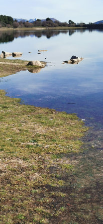 Calm lakeside view on a clear day with gentle ripples reflecting surrounding rocks and grassy shoreの写真素材