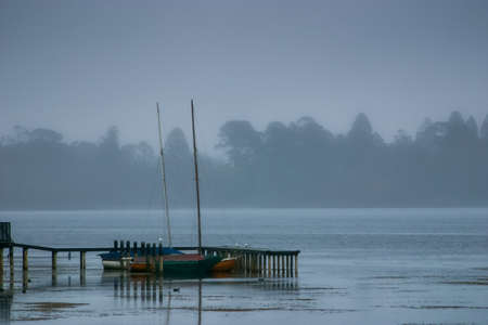 A cold foggy morning on the shore of Lake Wendouree in Ballarat, Victoria, Australia. Small sail boats moored alongside an old wooden pier strutting out into the shallow waters.の写真素材