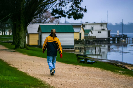 Lady wearing blue jeans, blue and yellow jacket, scarf and red gloves walking along gravel track around the shoreline of Lake Wendouree, Ballarat on a cold misty morning next to old wooden boat shedsの写真素材