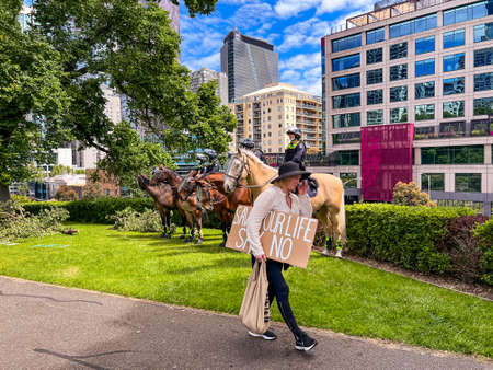 Melbourne, Victoria Australia - November 20 2021: Flagstaff Gardens Park A woman in a black hat carries her peaceful protest sign for freedom saying, save your life say no, in front of mounted policeのeditorial素材