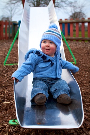 10 month old baby boy sitting at end of slide in play park in winter wearing blue coat and bobble hat. Happy and smilingの写真素材