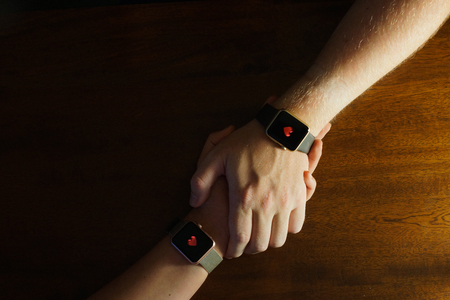 A couple hold hands whilst with hearts on their smart watches against a wooden background.の写真素材