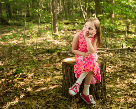 Young girl sitting on a stump in the forest, smiling and looking towards left side of frame and light.  Soft focus on background to allow for copy.  Dappled light coming through the trees for a soft, vintage feel.の写真素材