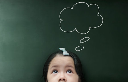 Cute thinking kid girl  with empty bubble looking on the blackboard background.の写真素材